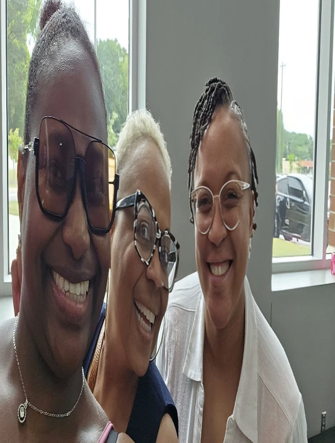 Three women smiling together indoors near a window.