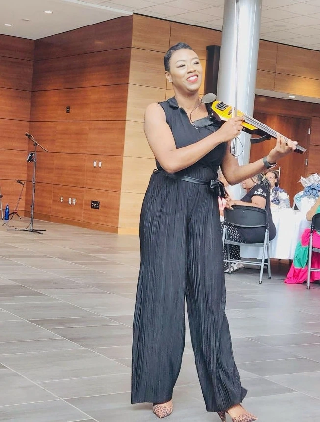 Woman playing a violin indoors in a black sleeveless jumpsuit.