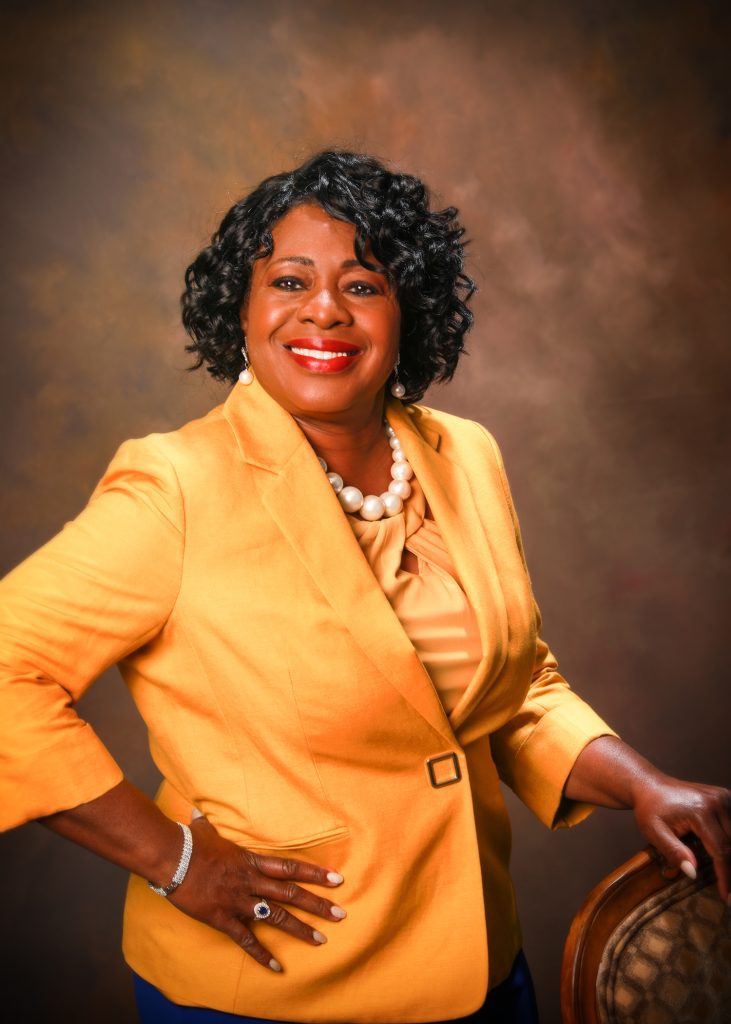 Confident woman in a yellow blazer smiling against a brown background.