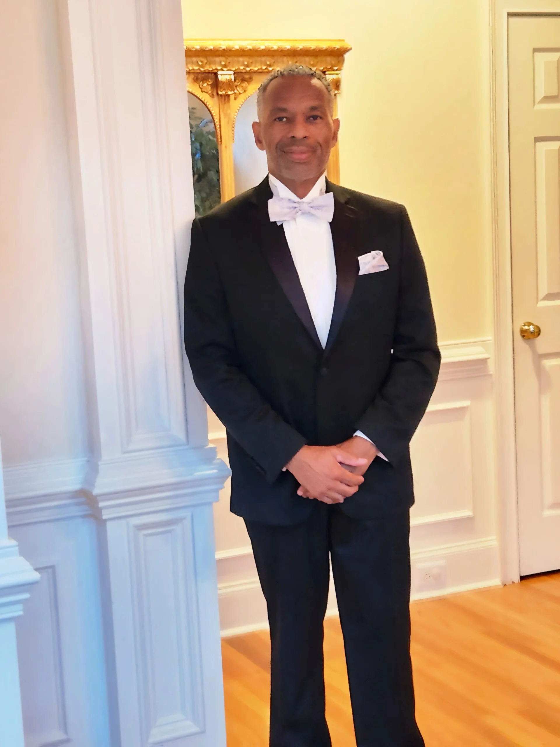 Man in a black tuxedo with a white bow tie standing indoors.
