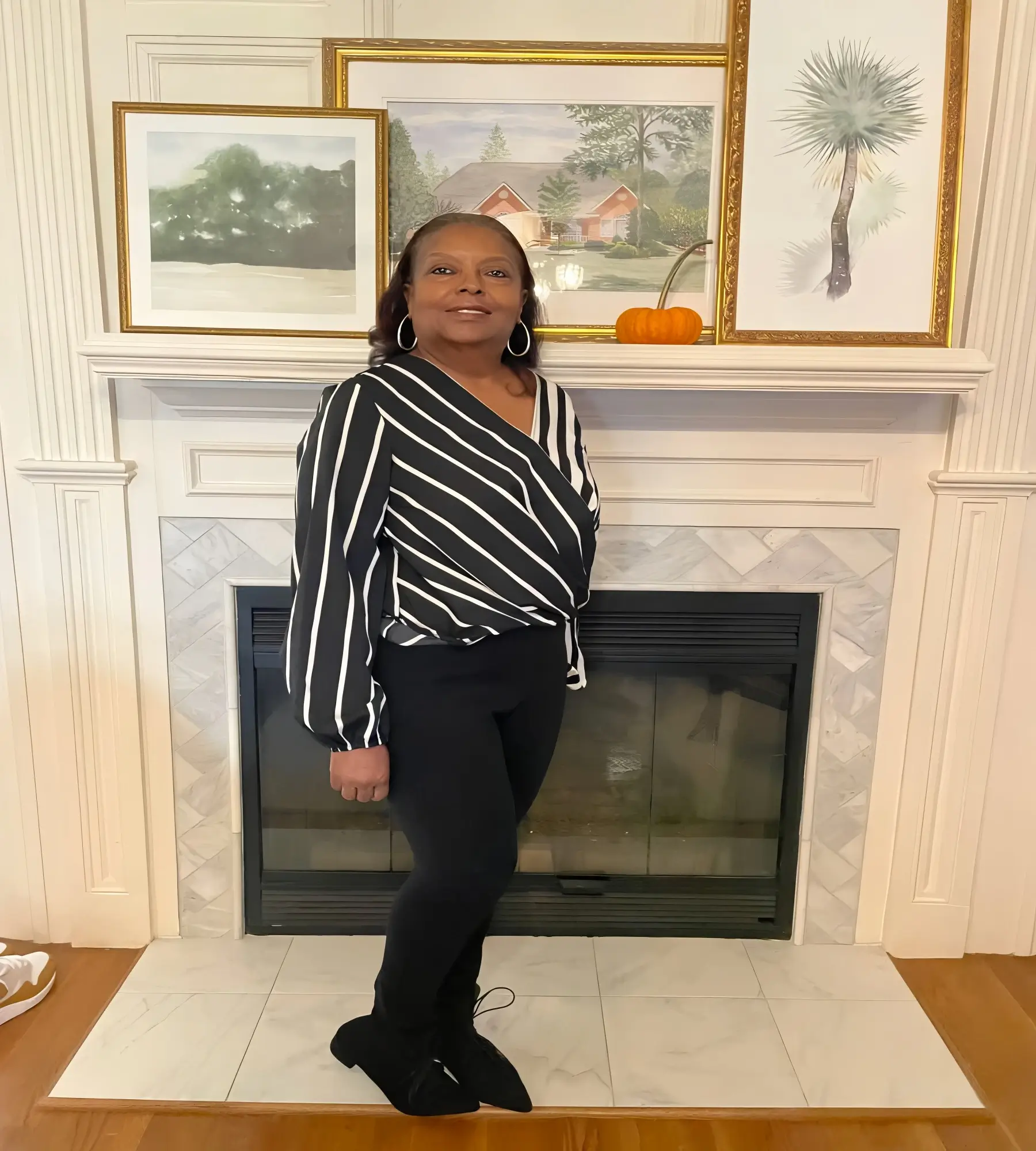 Woman posing confidently in front of a fireplace with framed artwork above.